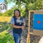 A woman holding two guns standing next to an archery target.